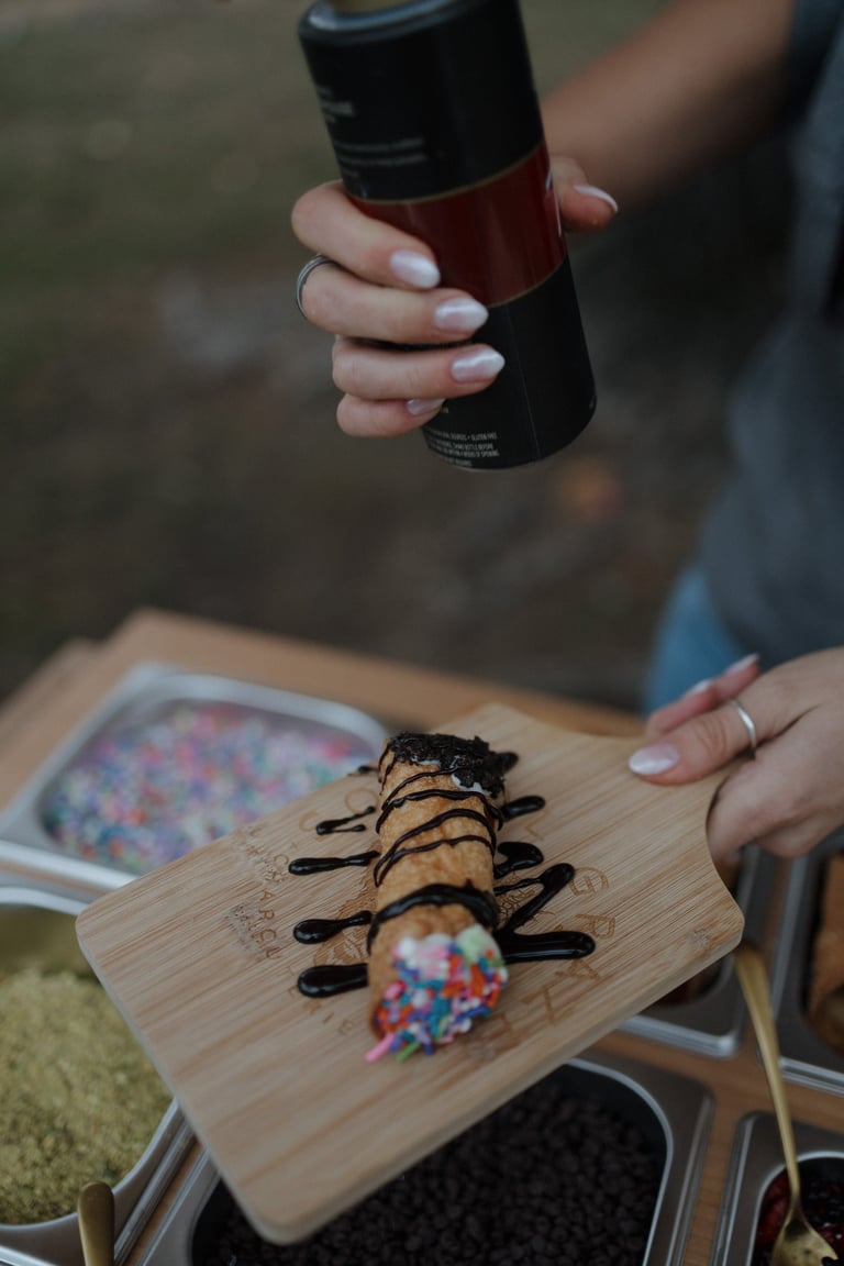 Person decorating a dessert with black drizzle sauce on wooden board, holding spray bottle