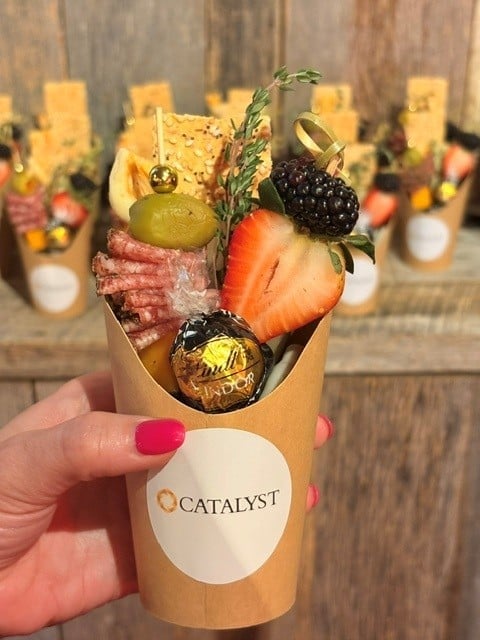 Hand holding a kraft paper cup labeled Catalyst filled with cured meats, olives, berries, and fresh herbs, with blurred similar cups in background