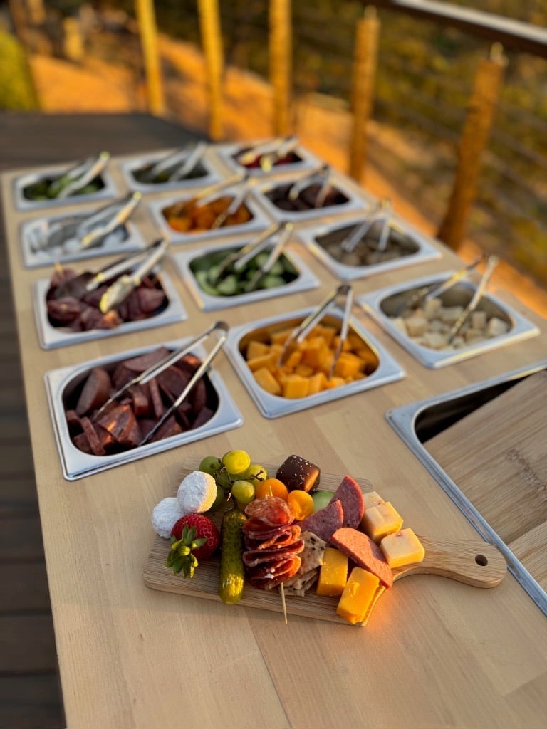 Charcuterie board display with cured meats, cheeses, and pickled vegetables on wooden surface with ingredient bowls arranged behind it