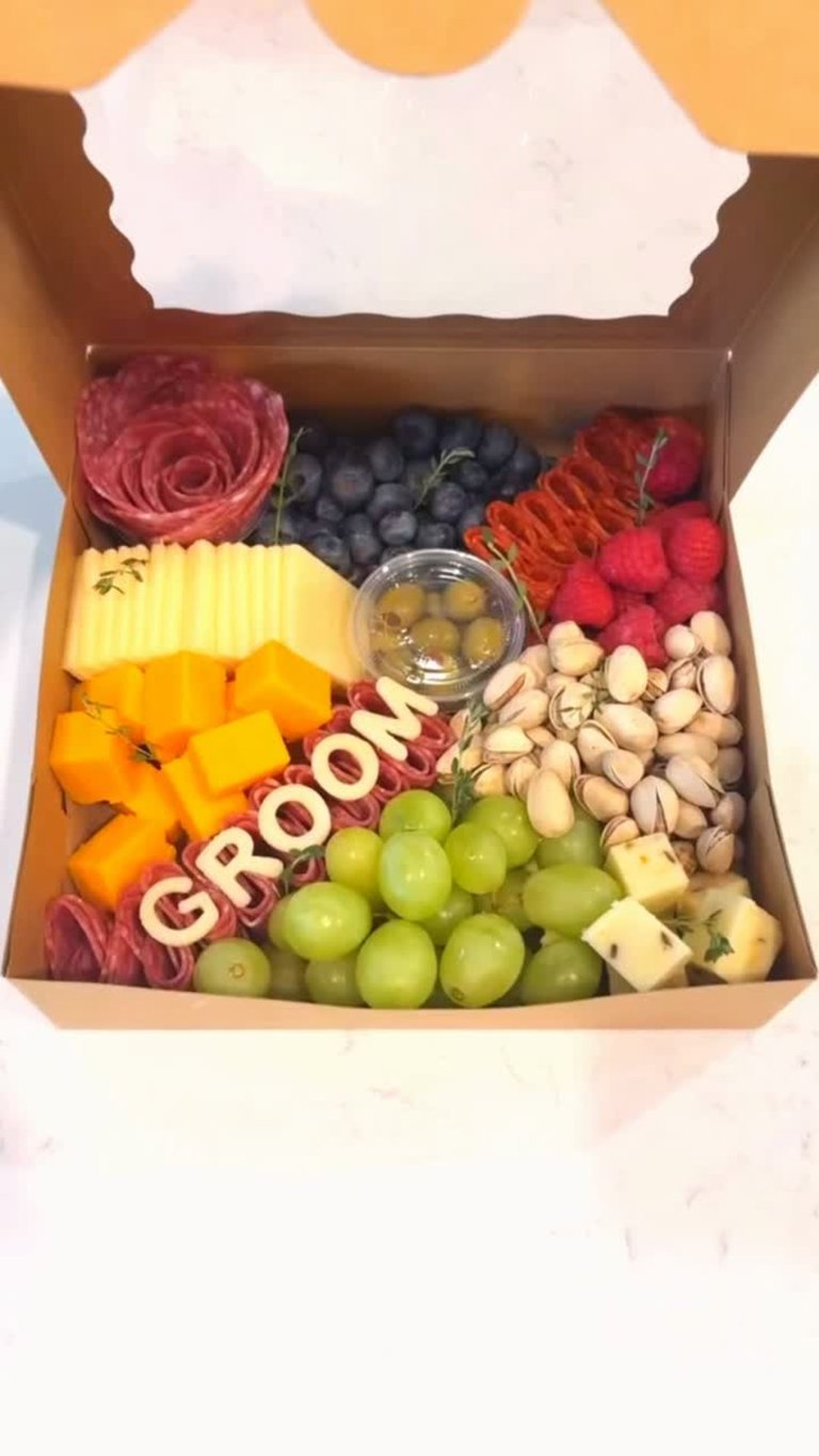 Open gift box with assorted fruits, cheeses, nuts, and a red rose arranged in sections including berries, grapes, citrus, and pistachios