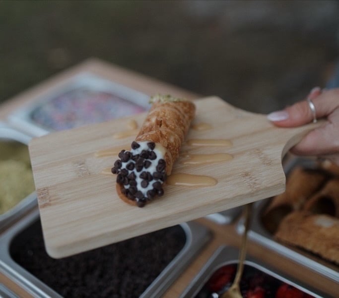 Cannoli with chocolate chips on wooden board held by hand over kitchen scale
