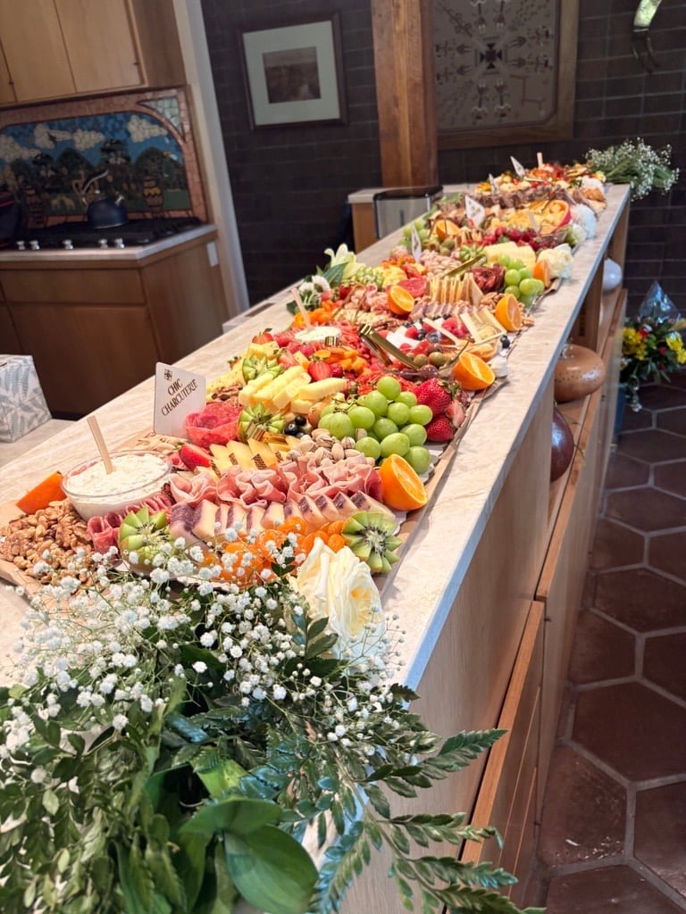 Long elegantly arranged charcuterie and fruit display on white tablecloths with fresh flowers and greenery in front