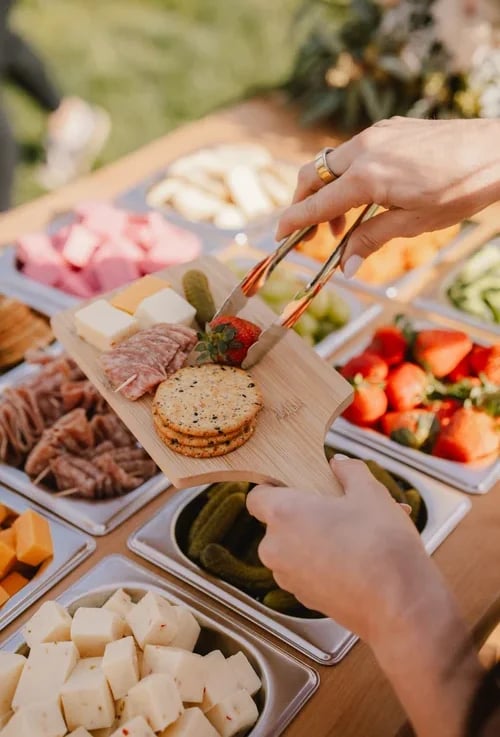 Person assembling a charcuterie board with cured meats, cheese, crackers, and fresh strawberries at an outdoor gathering