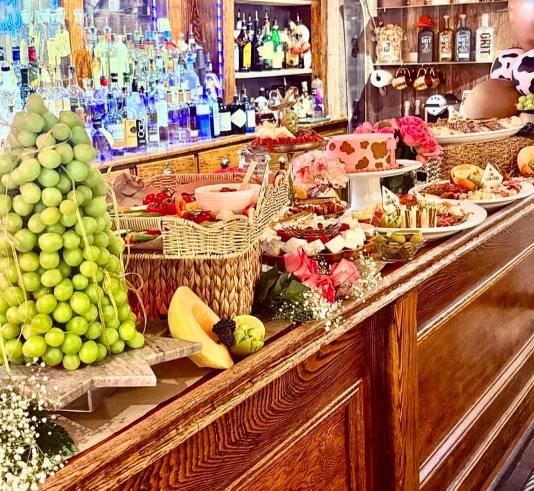 Upscale bakery and pastry shop display with pyramid of green apples, fresh fruit, flowers, and assorted pastries on wooden counter