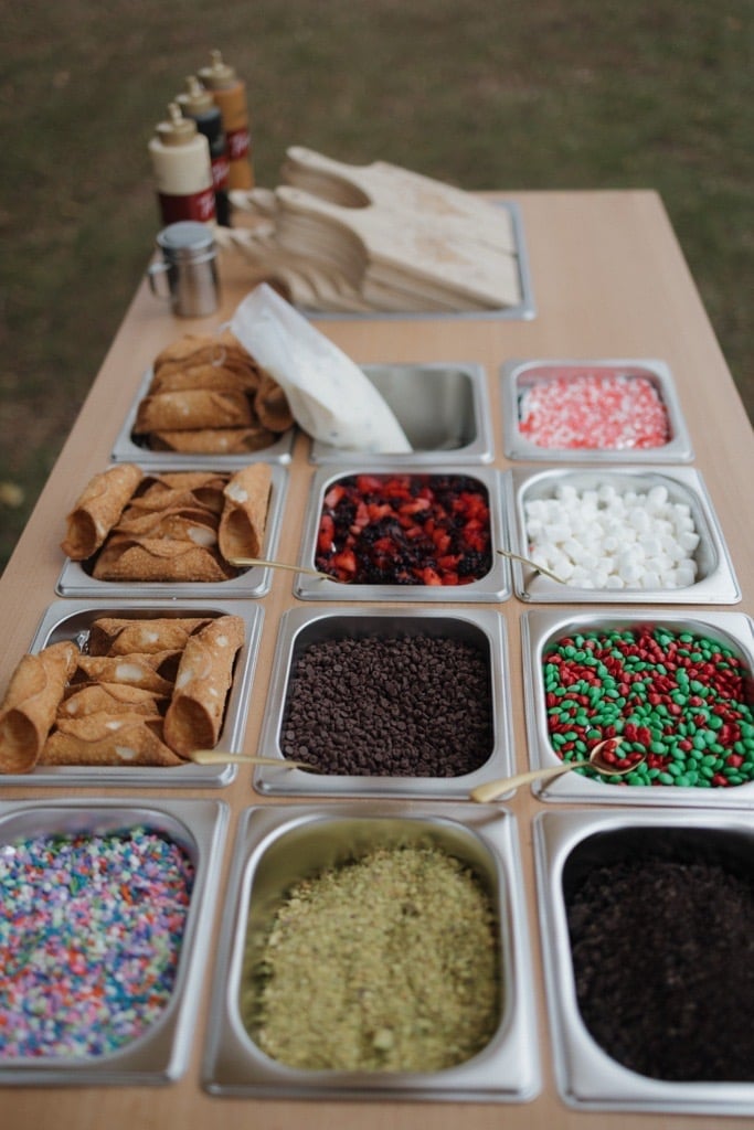 Overhead view of baking and decorating ingredients in metal containers, including cookies, sprinkles, berries, chocolate chips, and various toppings on a cardboard table