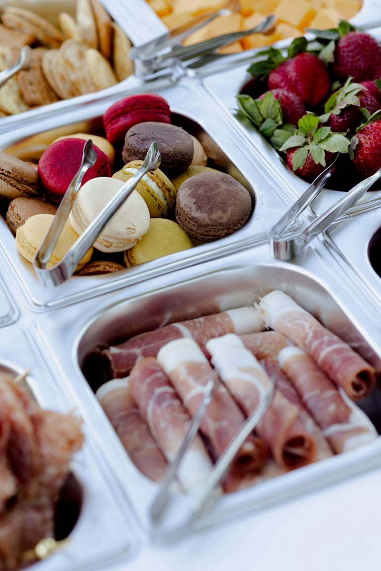 Overhead view of a charcuterie board with cured meats, fresh raspberries, macarons, nuts, and assorted pastries on white serving trays.