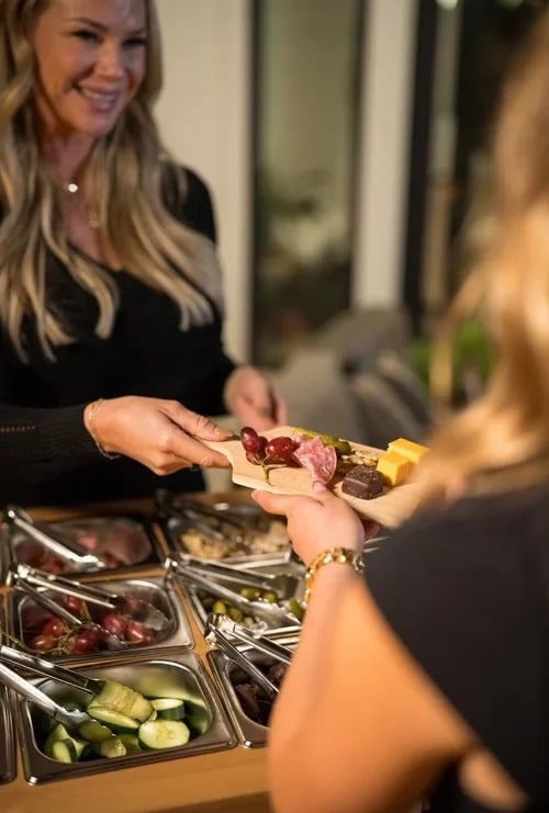 Woman serving charcuterie board items to another person at an event with appetizers visible