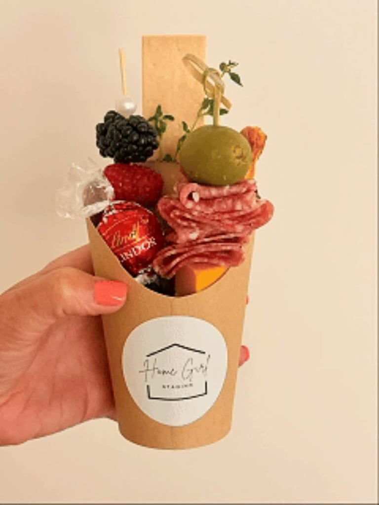 Hand holding a kraft paper cone filled with cured meats, fresh fruits, nuts, and herbs with a Home Staged logo label