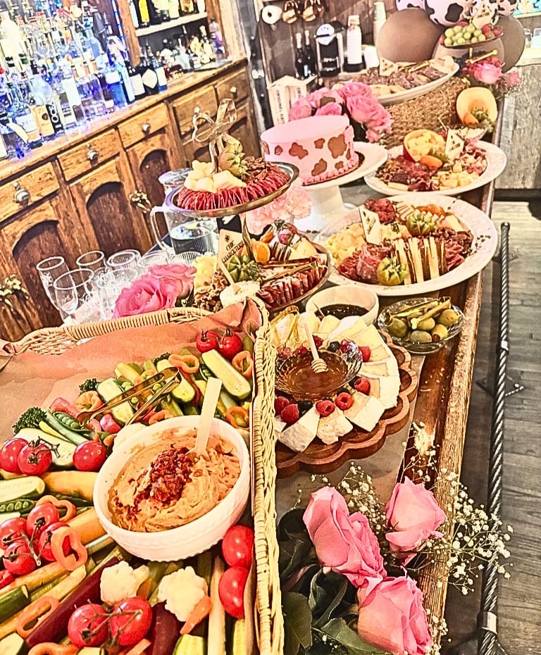 Elegant buffet display with gourmet appetizers, meats, cheeses, and cakes decorated with pink flowers and fresh produce on white platters